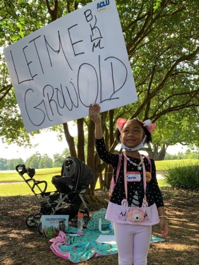 A young Black girl holding a sign reading "Let Me Grow Old"