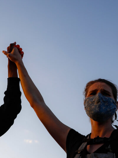 A black protester and a white protester hold hands while holding a sign that reads "We Can't Breathe"