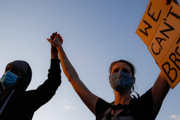 A black protester and a white protester hold hands while holding a sign that reads "We Can't Breathe"