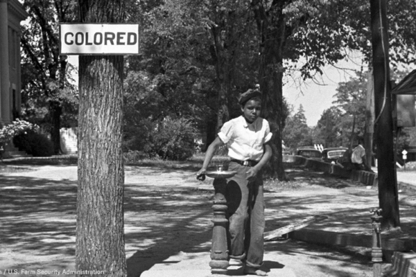 A Black boy stands next to a water fountain marked "colored" in this historical photo