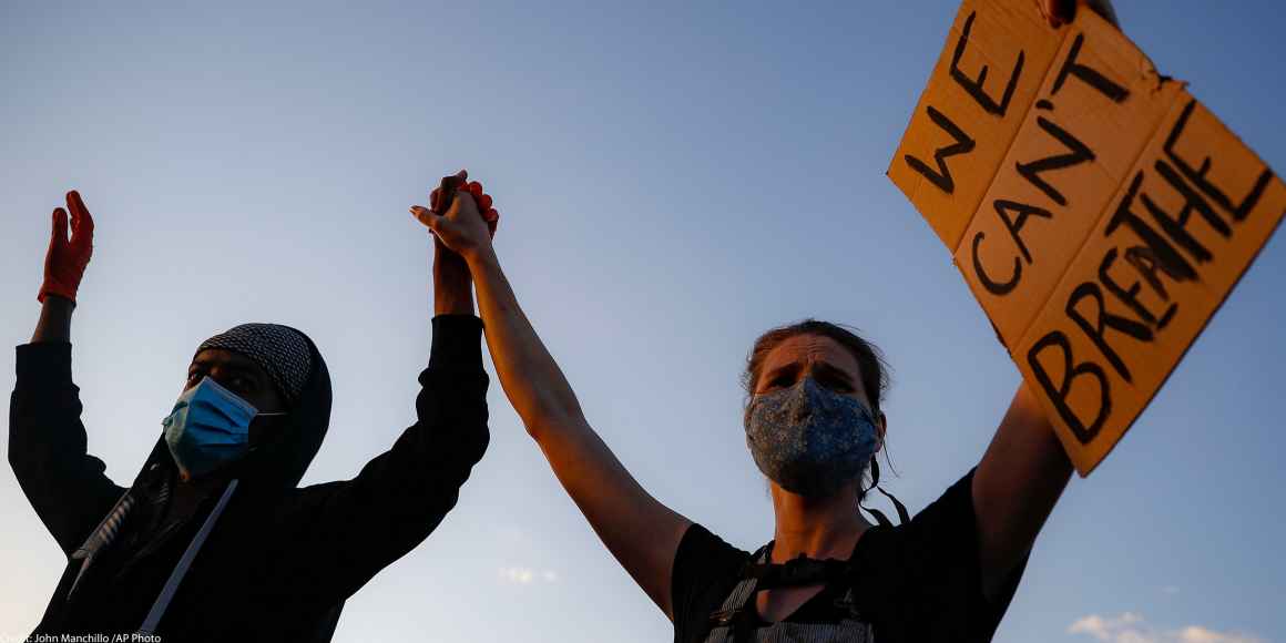 A black protester and a white protester hold hands while holding a sign that reads "We Can't Breathe"